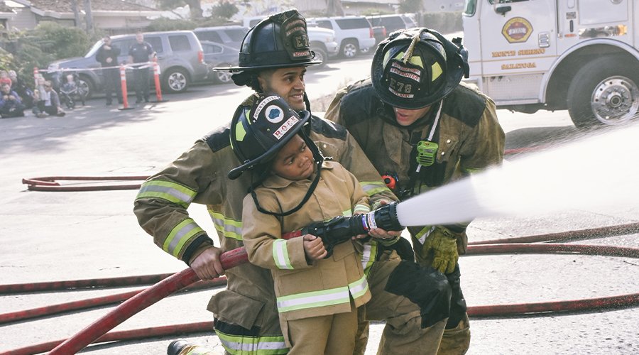 Firefighters showing child, in firefighter gear, how to use fire hose.